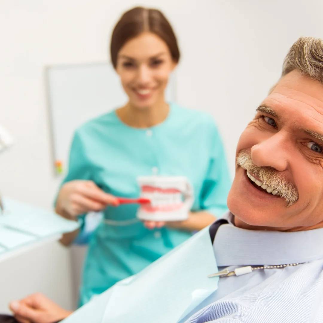 Middle-aged man smiling in dental clinic with female dentist holding tooth model in background